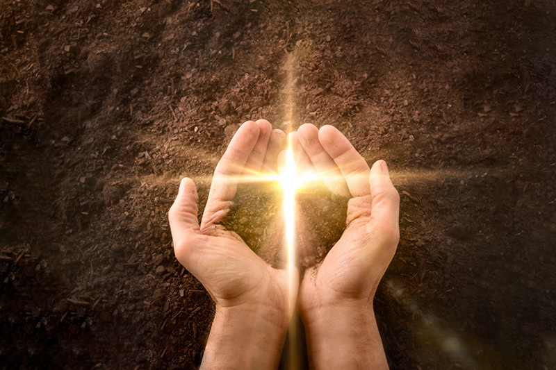 Hands together with a small mound of earth with a golden flash in the shape of a cross on a background of earth. Jesus resurrection concept. Horizontal composition. Top view.
